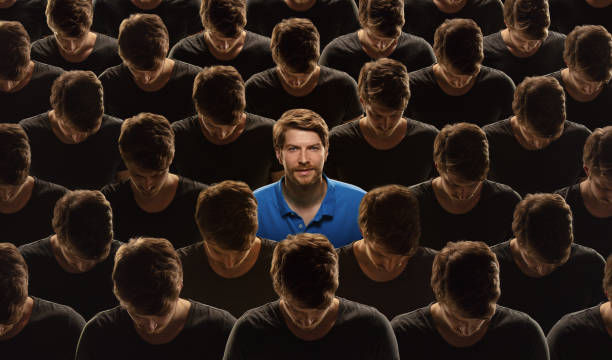 Man in blue shirt stands amongst identical figures in black shirts with bowed heads, creating a striking contrast. Mood of individuality.