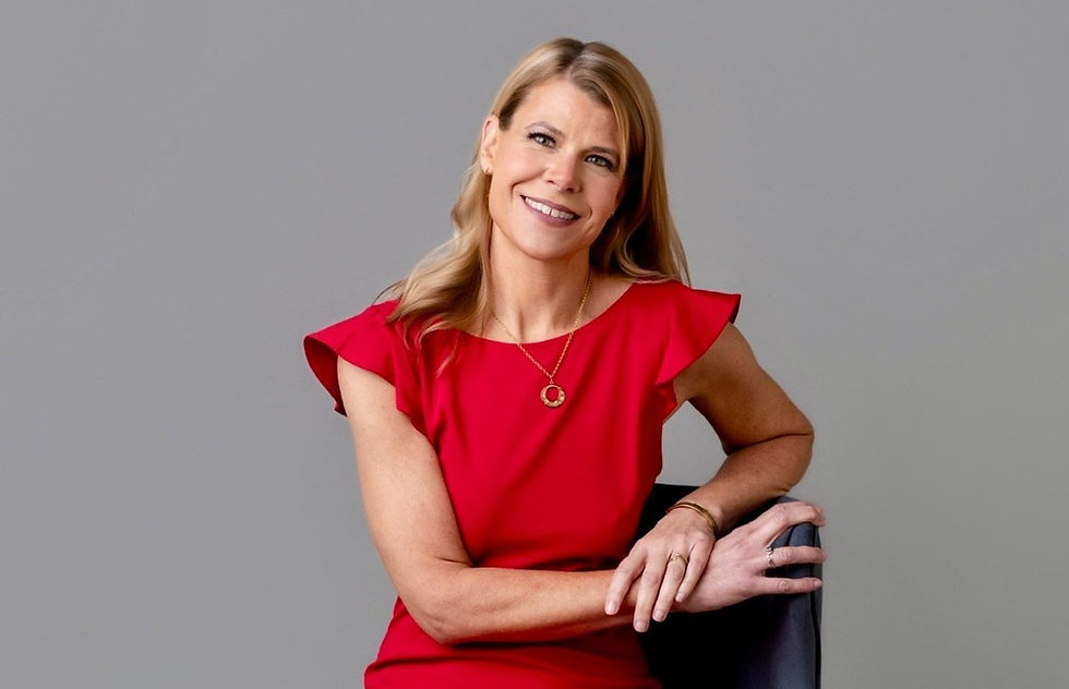 Smiling woman in a red dress with a necklace, seated against a gray background. Relaxed pose, blonde hair, joyful expression.