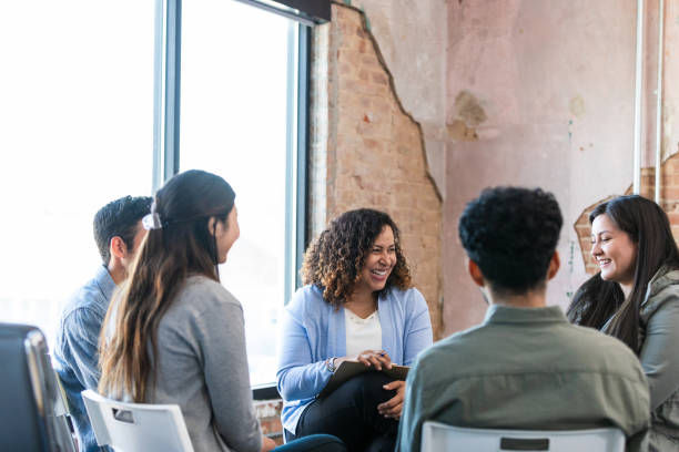 Group of diverse people seated in a circle, talking and laughing. Exposed brick wall and window in background, creating a casual, positive mood.