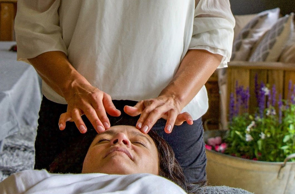 Woman giving a facial massage to a relaxed person lying down. Cozy setting with pillows and purple flowers in the background.