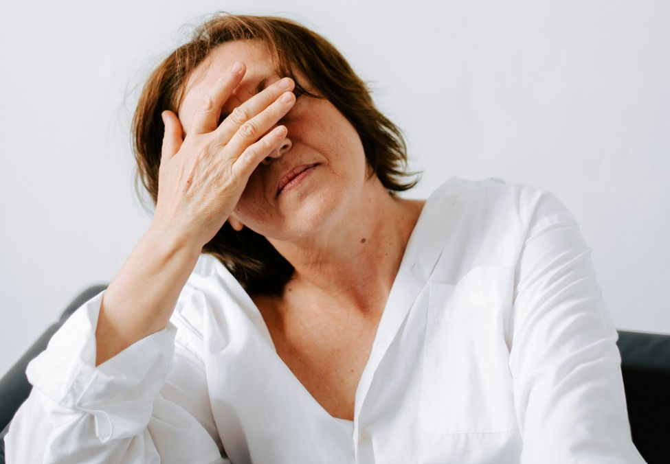 Woman in a white shirt covers her face with one hand, looking tired or stressed against a plain white background.