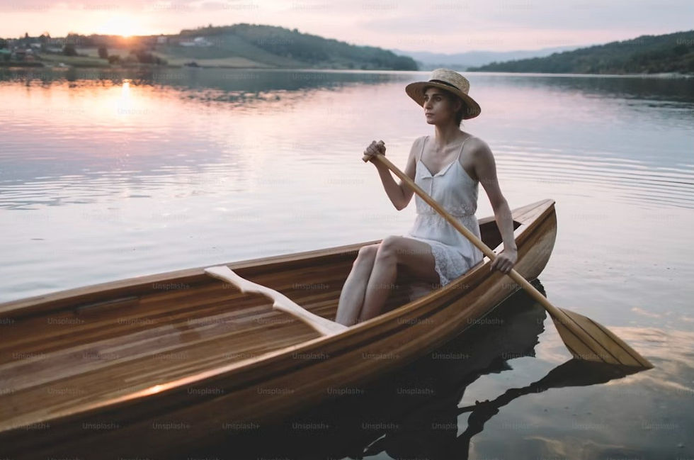 Woman in a straw hat rows a wooden boat on a calm lake at sunset, with pink reflections and distant hills.