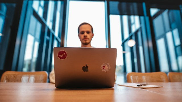 Man works on a sticker-covered laptop at a wooden table in a glass-walled office, with a notepad beside him.
