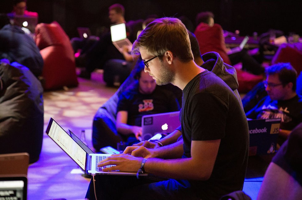 Young man with glasses types on a laptop at a dim, purple-lit coding event, with other people working on laptops behind him.