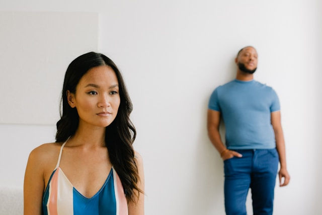 Woman in a striped dress stands in foreground with a serious look; a man in blue leans against a white wall behind her.