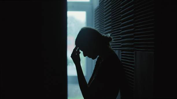 Silhouette of a person in profile holding their forehead, standing in a dimly lit hallway. The mood appears contemplative and somber.