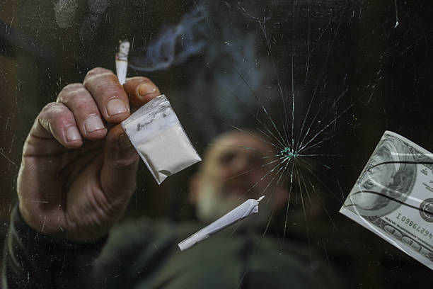 Hand holds a small white packet and cigarette near a cracked glass. A dollar bill floats. Smoky atmosphere, dark background, tense mood.