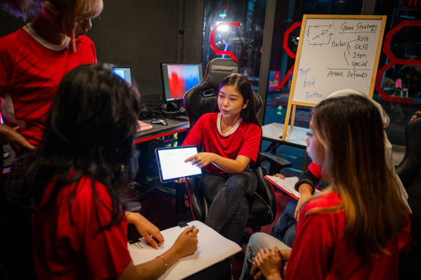 A group of people in red shirts discuss gaming strategies in a room with computers. One holds a tablet, and a board shows game notes.