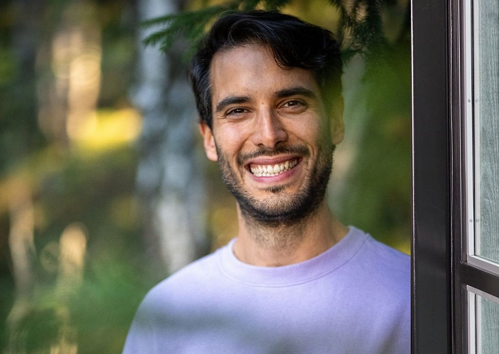 Smiling man in a lavender shirt stands by a window, with a forest background. Sunlight filters through green leaves, creating a warm atmosphere.