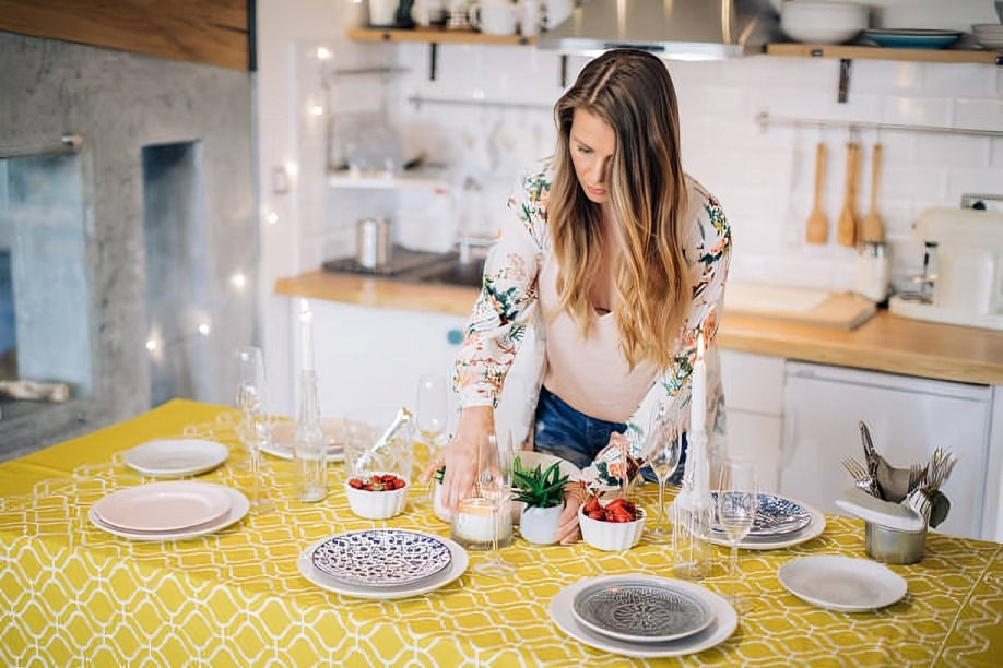 Woman setting a table with candles, plates, and strawberries on a yellow cloth. Kitchen background, cozy lighting, floral shirt.