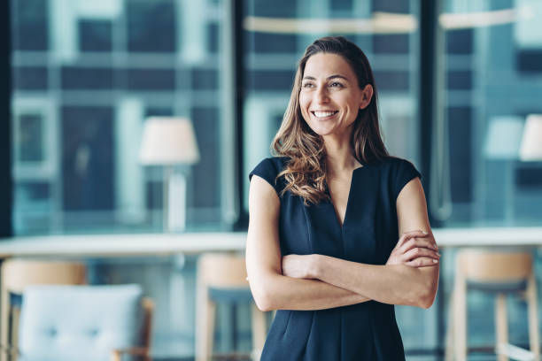 Smiling woman in a black dress stands with arms crossed in a modern office, with blurred windows and chairs behind her.
