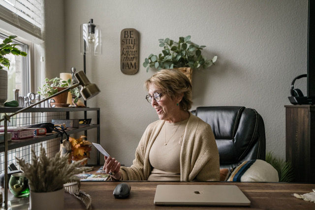 Older woman reads a paper at a desk in a cozy home office, with plants, a laptop, and a wall sign reading Be still and know that I am.