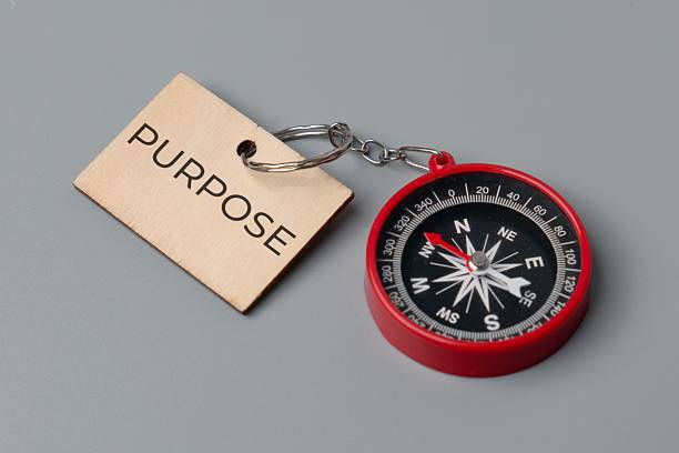 Red compass with a brown tag labeled "PURPOSE" on a gray surface. The image conveys a sense of direction and intention.