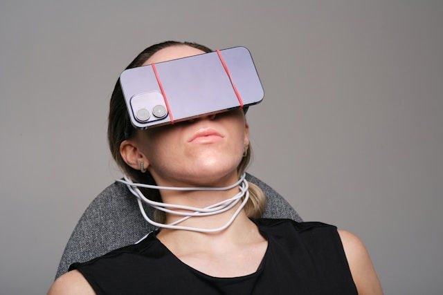 A person leans back in a chair with a phone strapped to their face using rubber bands. White cables are wrapped around their neck. Neutral background.