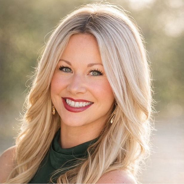Smiling woman with long blonde hair in a green top outdoors. Bright sunlight creates a soft, warm background, enhancing her cheerful expression.
