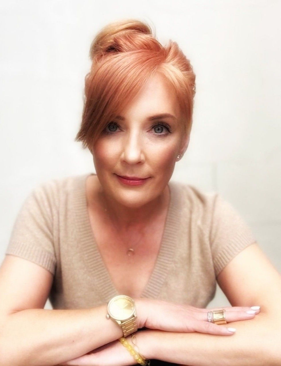 Woman with red hair and fair skin, smiling softly, wearing a beige top and gold watch, set against a plain white background.