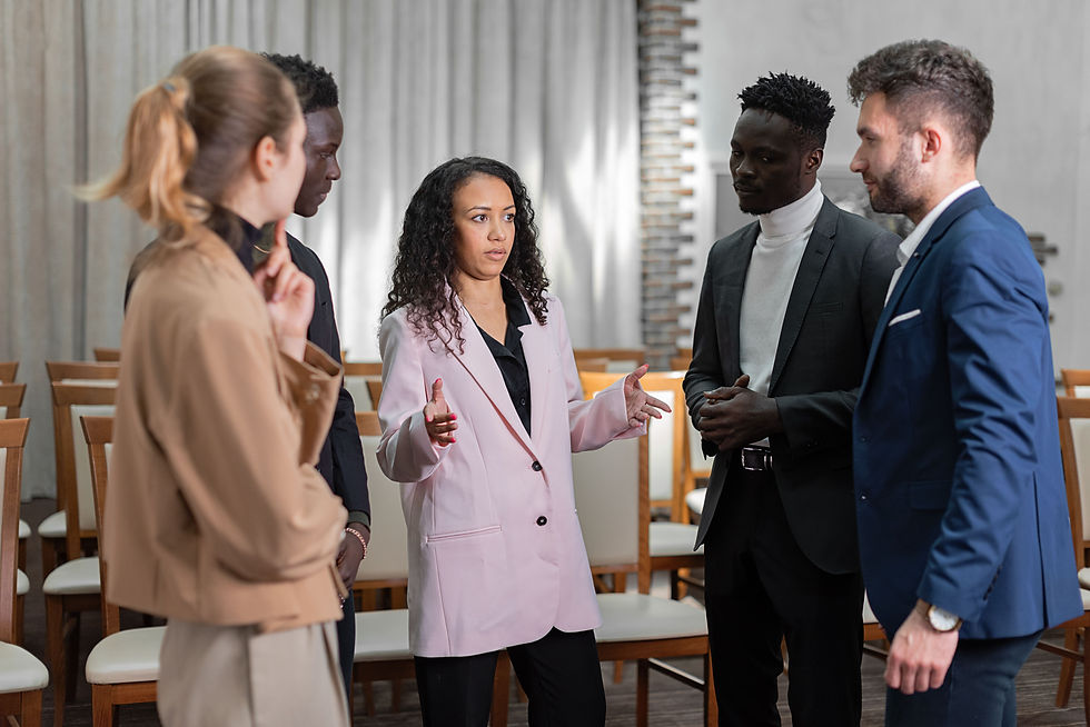 Five business professionals stand in a meeting room, listening to a woman in a pink blazer speaking with hands raised.