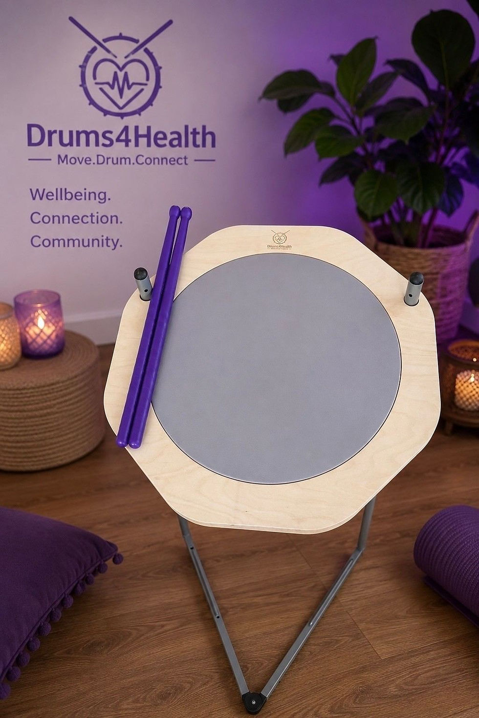 Purple-lit Drums4Health drum setup with sticks, candles, plant, and wall text: Wellbeing. Connection. Community.