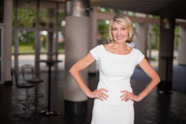 Smiling blonde woman in a white dress stands hands on hips in a modern lobby with blurred tables and windows.