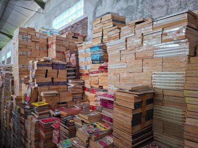 Stacks of books piled high in a warehouse-like room, with colorful covers among mostly brown papered bundles, creating a crowded scene