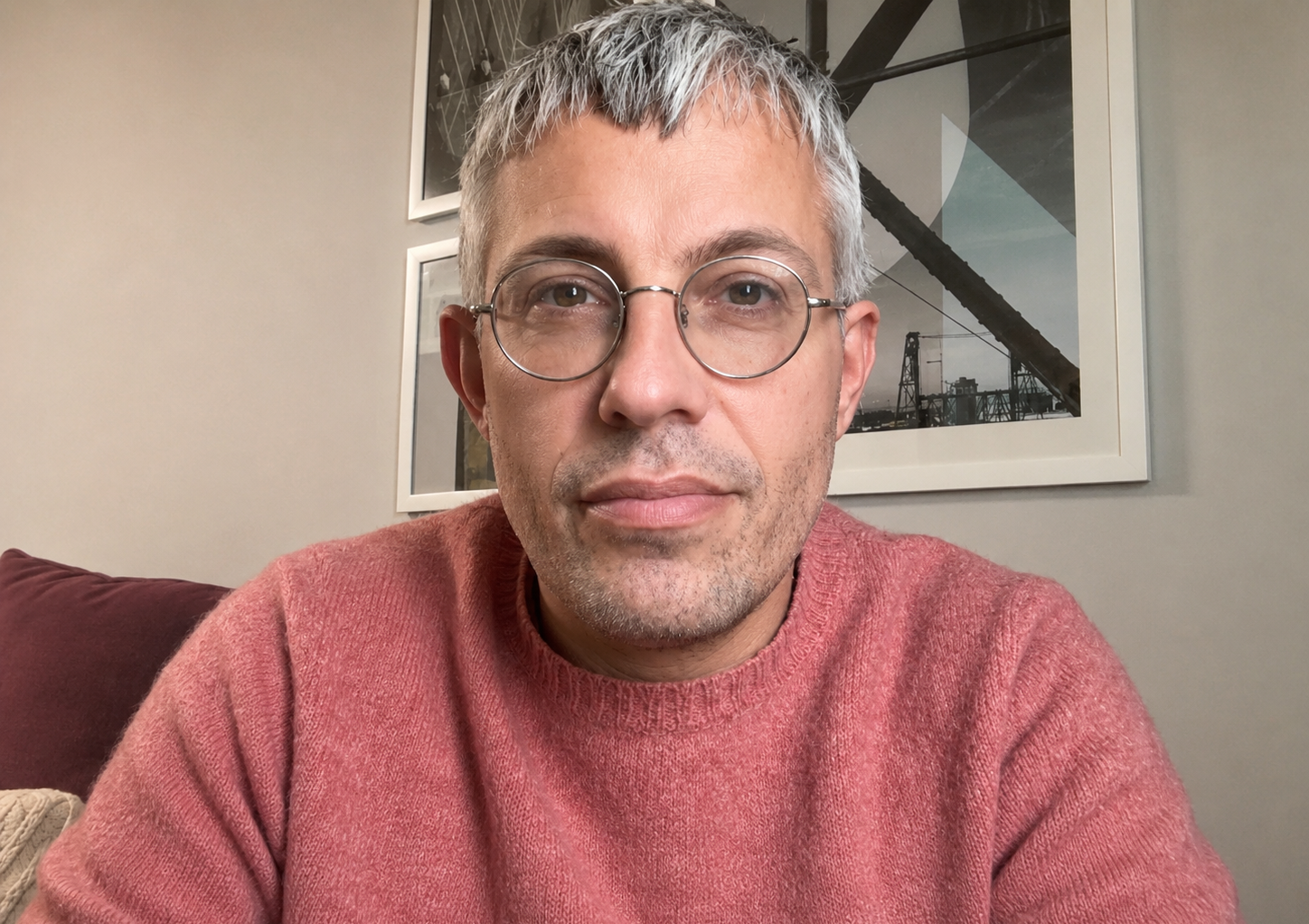 Close-up selfie of a man with gray hair and glasses in a pink sweater, sitting indoors with framed art behind him.
