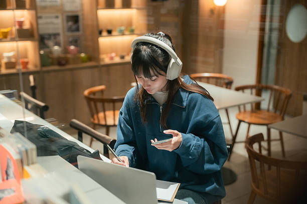 Young woman with headphones writes in a notebook while checking her phone at a warm, empty café table.