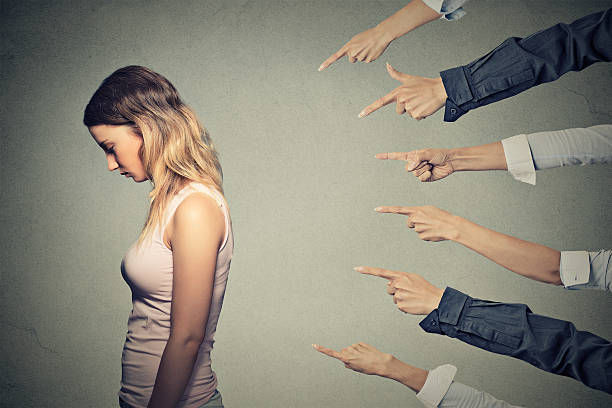 Sad woman looks down while several pointing hands accuse her against a plain gray background.