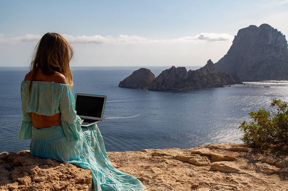 Woman in a flowy teal dress sits on a cliff, using a laptop while gazing at the ocean and islands in the distance under a clear blue sky.