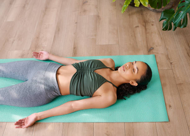 Woman lying on a green yoga mat in a relaxed pose, eyes closed. Wood floor and green plant in background. Calm and peaceful mood.