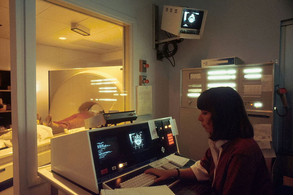 Technician operates a computer in a dim MRI control room. Patient in scanner behind glass. Monitors display medical images.