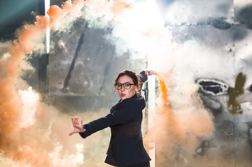 A woman in a black blazer and glasses poses dynamically amid swirling orange smoke, set against a metallic, industrial background.