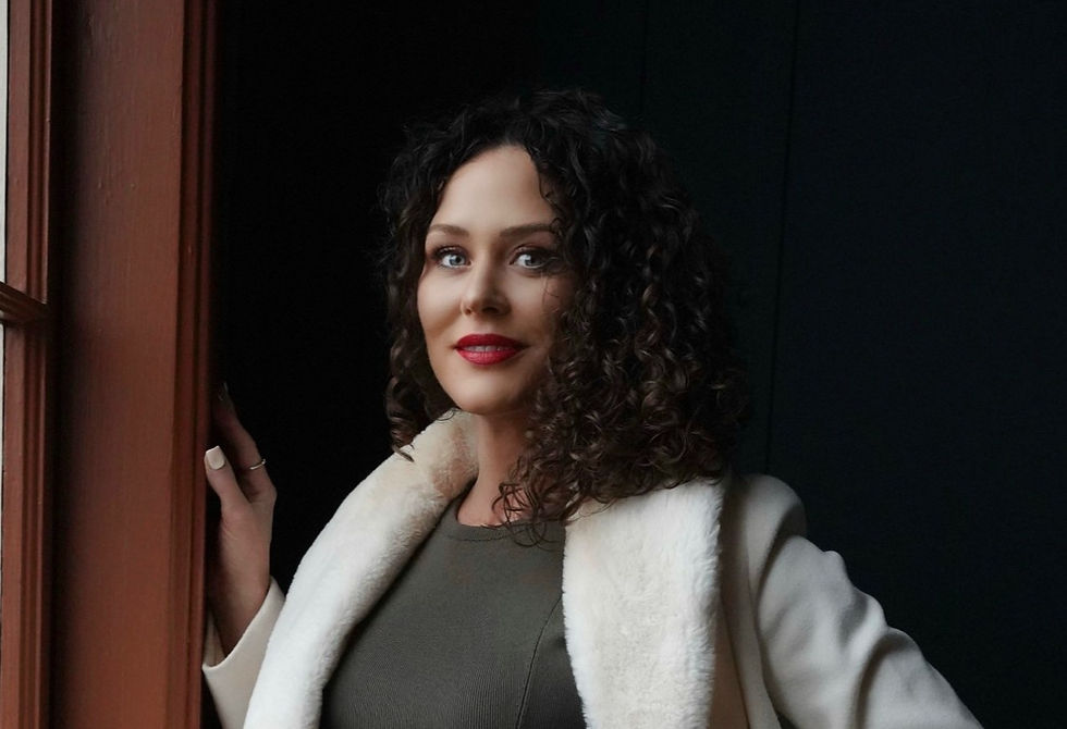 Woman with curly hair stands by a window wearing a white coat with faux fur collar, smiling. Background is dark, creating a warm mood.