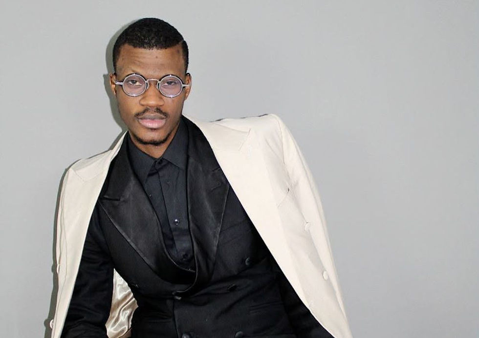 Stylish man in cream suit and black shirt adjusts glasses against a plain gray background, looking thoughtful.