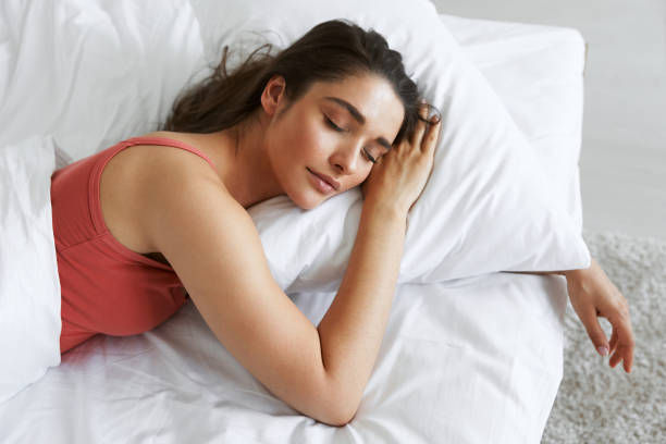 Woman in a pink top sleeping peacefully on white bedding. Bright, serene room with soft lighting, conveying calm and relaxation.