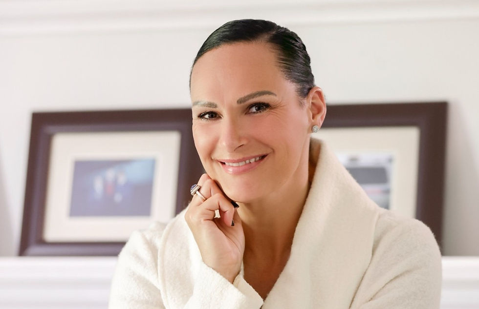 Smiling woman in a white coat poses indoors, hand on chin. Blurred framed pictures in the background. Warm, inviting mood.