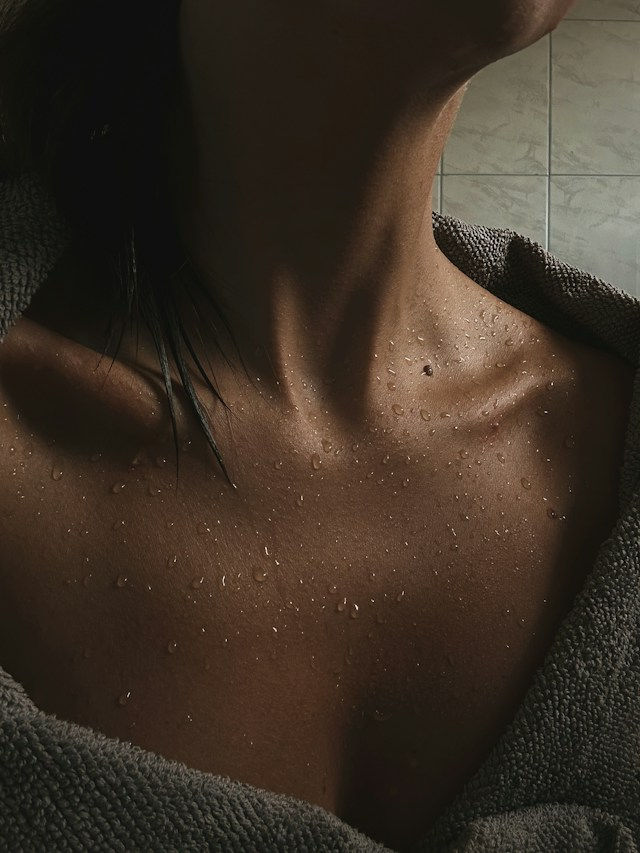 Close-up of a wet neck and chest wrapped in a gray towel, with bathroom tiles behind, creating a moody, intimate feel.
