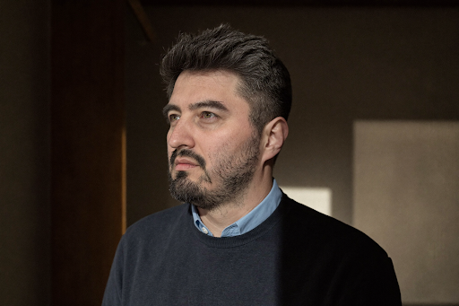 A man with dark hair and a beard looks contemplatively to the left, wearing a blue shirt and dark sweater, in a dimly lit room.