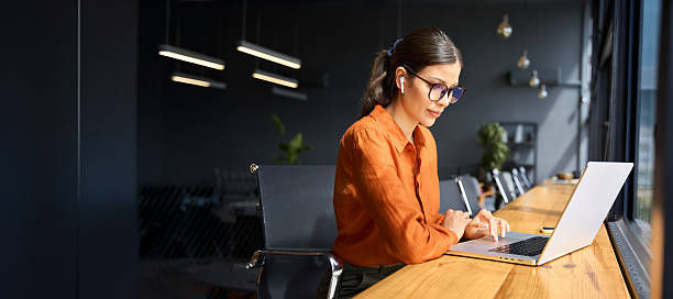 Woman in an orange blouse and glasses works on a laptop at a wooden table in a modern office, focused and intent.