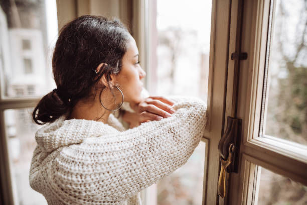 A woman in a cozy knit sweater gazes thoughtfully out of a window, resting her chin on her hands.