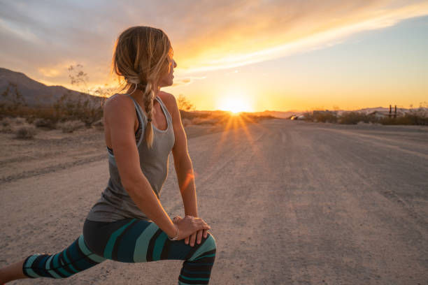 Woman doing a lunge on a dirt road at sunset. She wears a tank top and striped leggings. The sky is vibrant orange and serene.