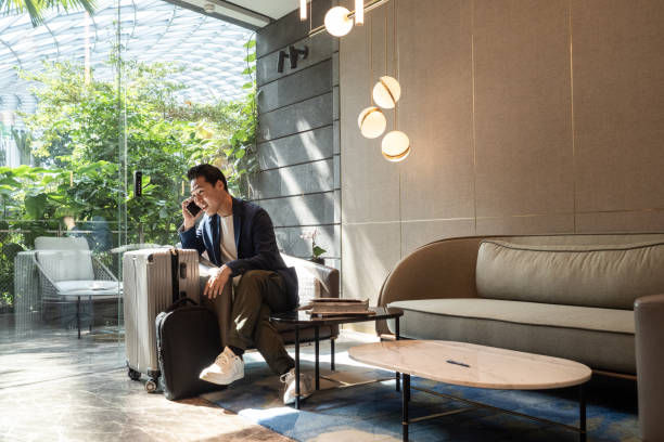 Man in suit talking on phone, sitting with luggage in a bright, modern lounge. Greenery outside glass wall, pendant lights above.