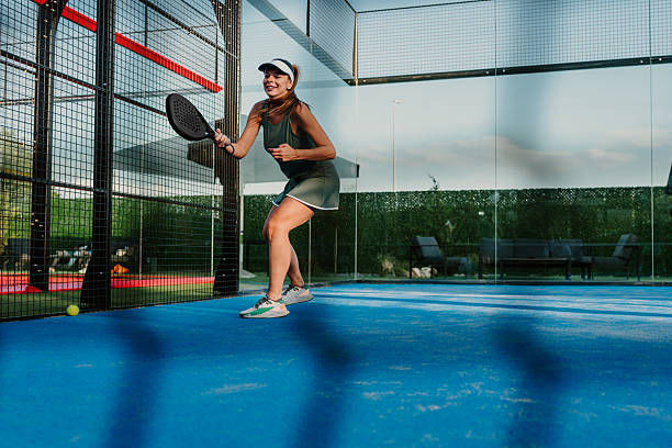 Smiling woman plays padel on a blue outdoor court, holding a paddle near the net with glass walls and greenery behind.