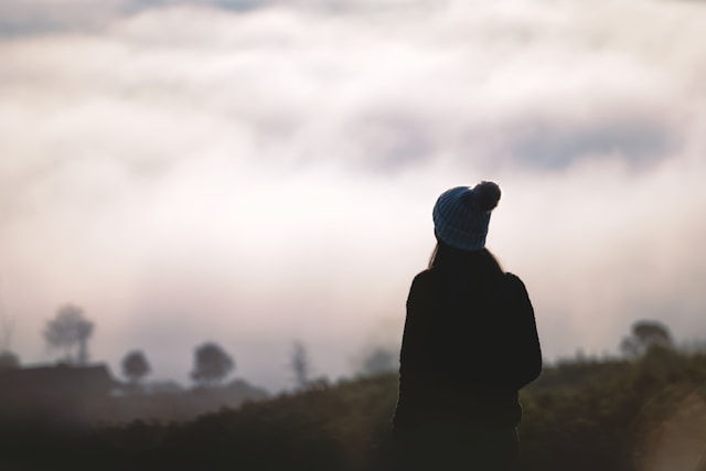 The photo shows a silhouette of a person wearing a beanie, gazing at a vast landscape covered in thick fog, creating a sense of mystery, solitude, and contemplation.