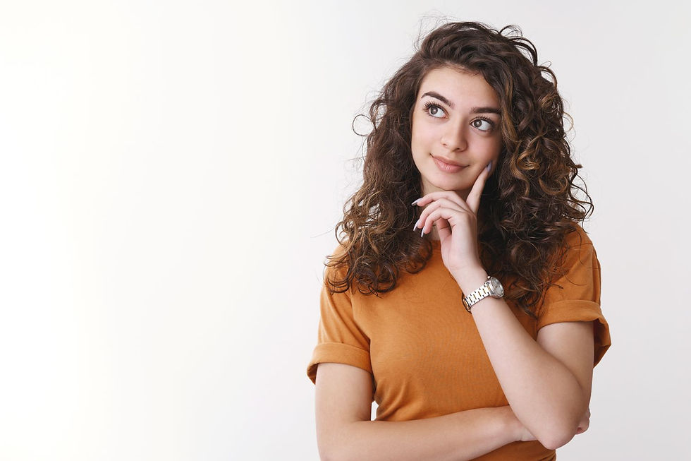 Young woman with curly hair in an orange shirt, thinking with a hand on her chin. Light background, thoughtful expression.