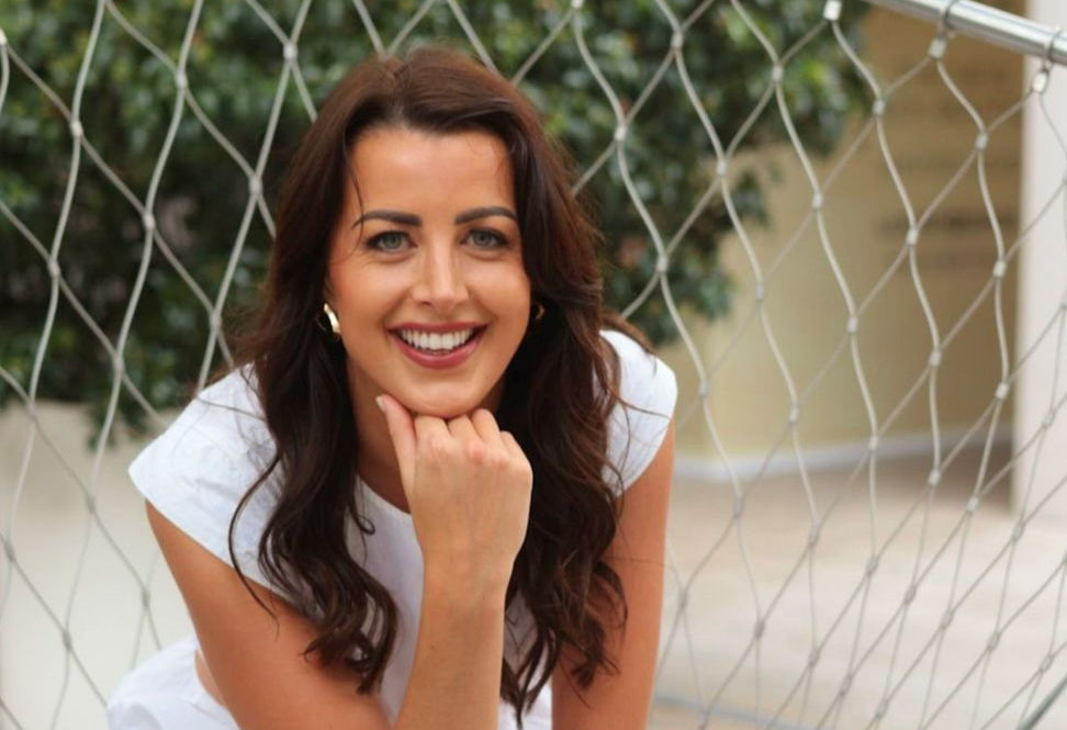 Woman smiling, leaning on a hand in front of a net fence. Blurred greenery in background, wearing white. Bright and cheerful mood.