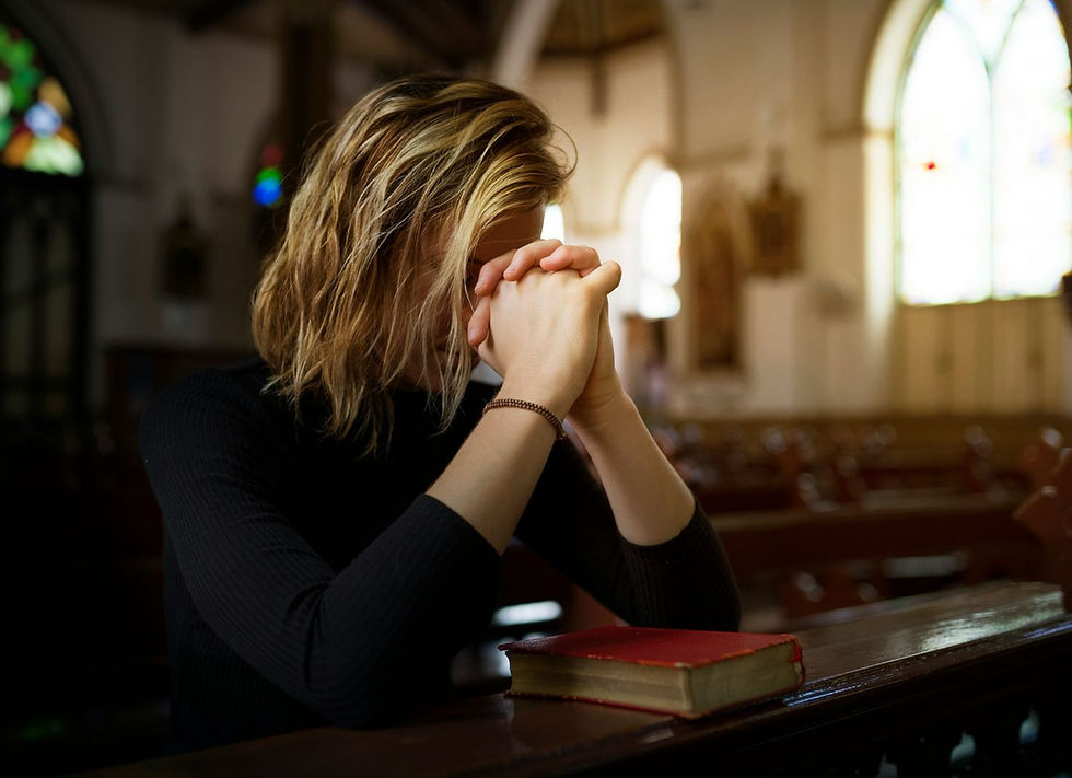 Person praying with folded hands in a church, sitting alone on a pew. A red book is nearby. Soft light filters through stained glass windows.