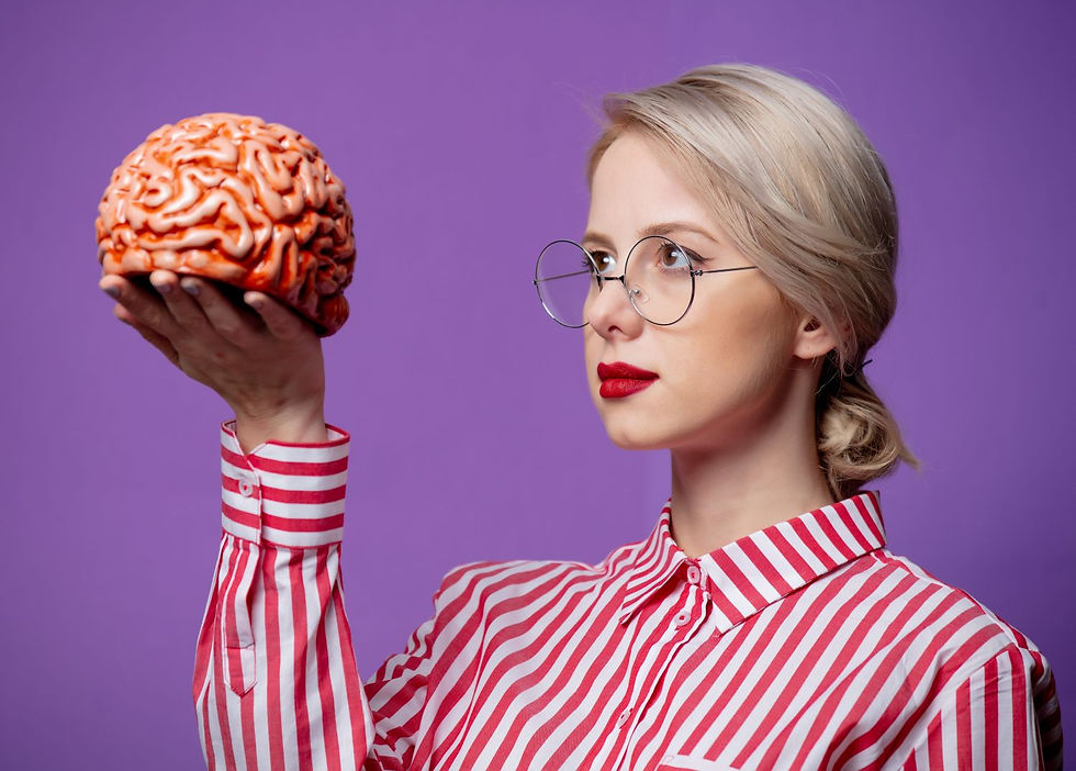 A woman wearing glasses and a red striped shirt is holding a model of a brain in her hand while looking at it thoughtfully against a purple background.