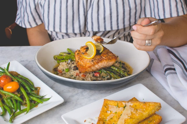 Person in a striped shirt eating salmon with lemon on rice and asparagus. Sides of green beans and cornbread on a marble table.