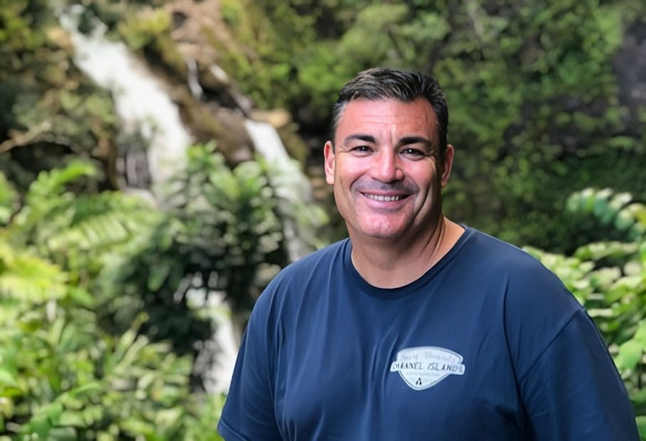 Smiling man in blue shirt with tropical print stands in front of a lush forest and waterfall, creating a serene outdoor setting.