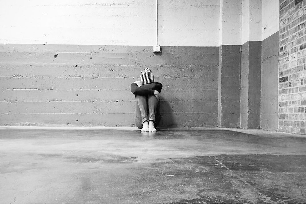A person sitting alone on the floor in a bare, industrial-style room, hugging their knees with their head down, conveying a sense of isolation or sadness.
